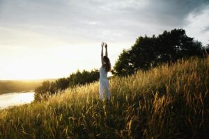 A woman in a white dress enjoys the sunrise in a scenic grassy field by the water.